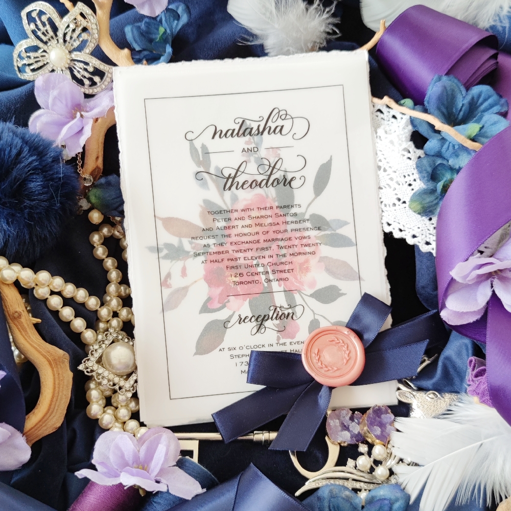 A two layer invitation with the invitation wording printed on a vellum page sitting on top of an ice pearl paper with bright pink and navy flowers printed.  Tied together with a navy bow and blush wax seal.