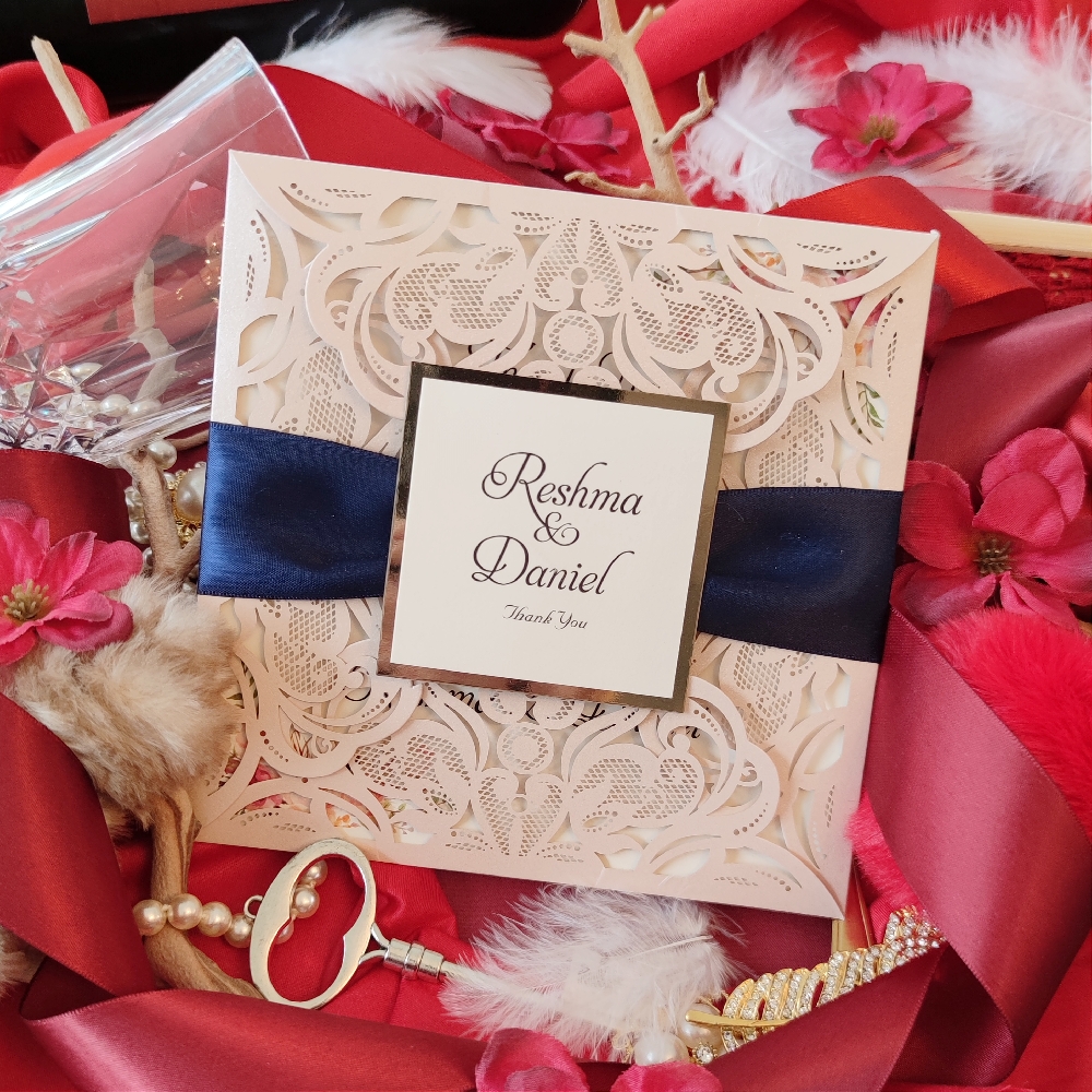 Blush lace cut wedding invite design with thick navy ribbon and layered square tag in the middle.  The lace design opens four ways to reveal the wedding invitation wording.