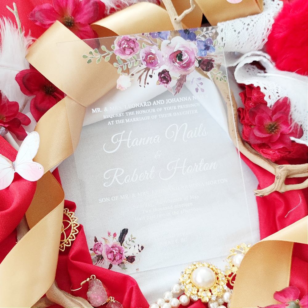 A clear acrylic wedding invitation with white ink and purple and lavender flowers.