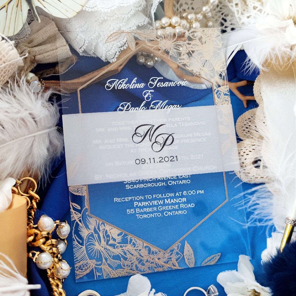 This is a clear acrylic wedding invite with a vellum belly band wrapped around the invite.  The main text is printed in a white UV ink.  There is a gold geometric border with floral designs surrounding the text.
