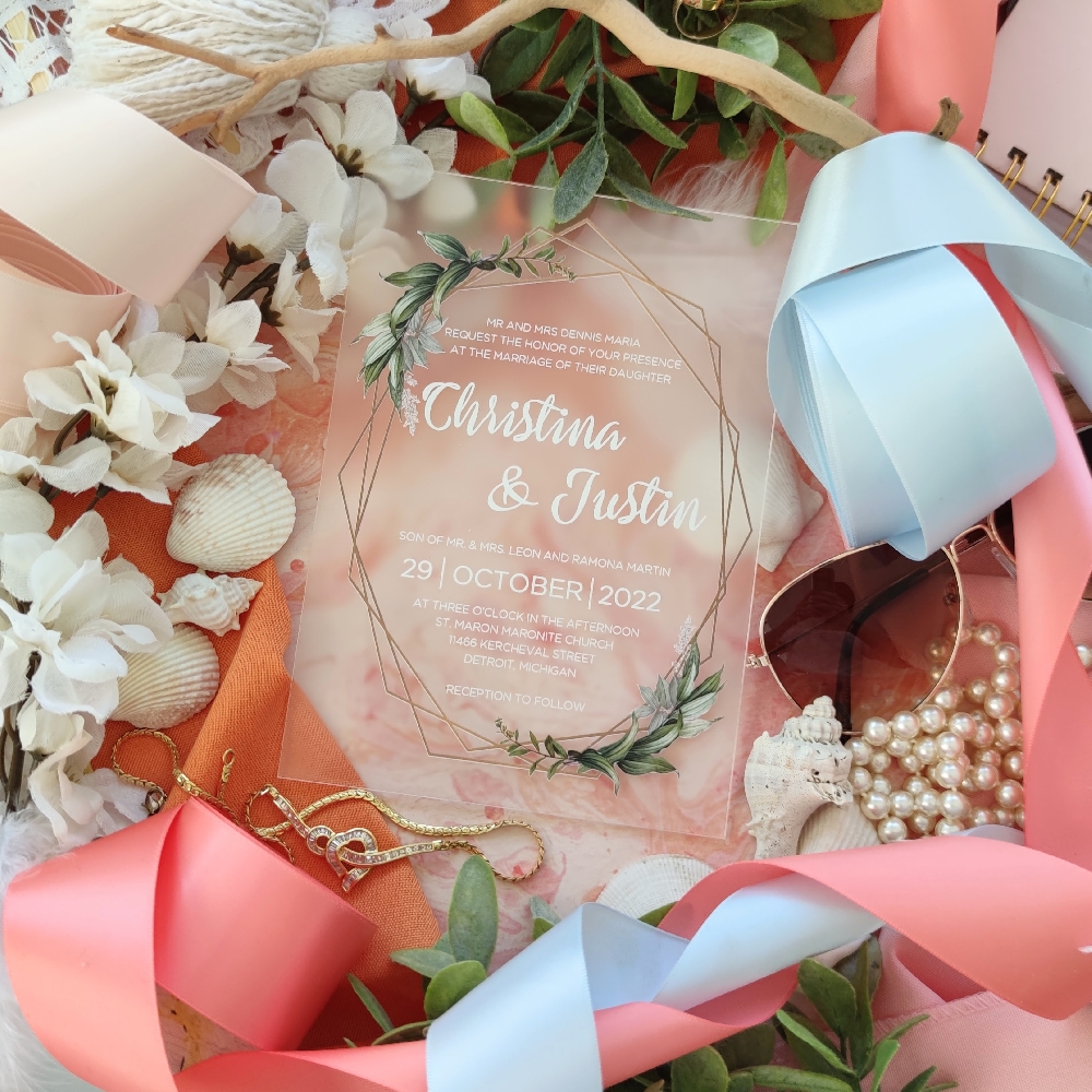 A frosted acrylic wedding invitation with white writing and greenery with geo-shape border.