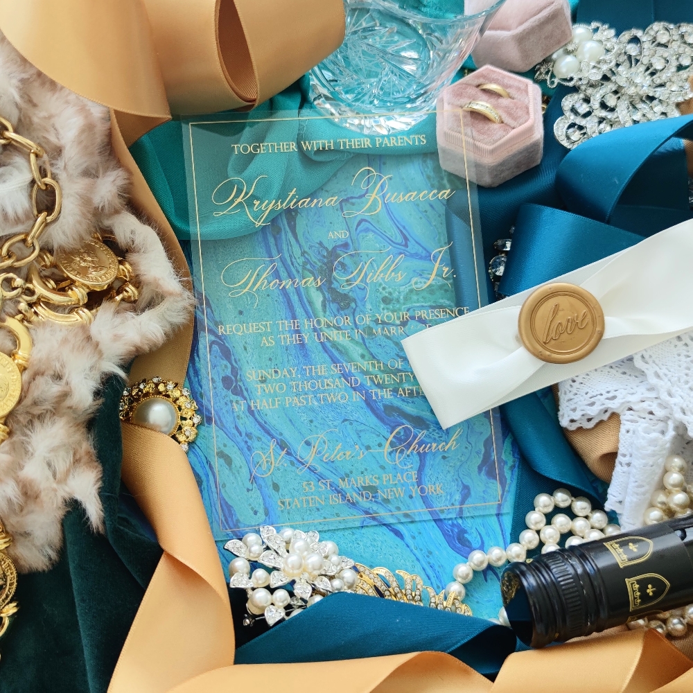 Clear acrylic wedding invitation with gold ink printing and an ivory ribbon and gold wax seal.