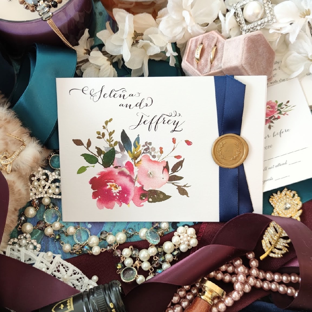 Folded wedding invitation with a navy ribbon and gold wax seal and pink floral bouquet printed on the cover.