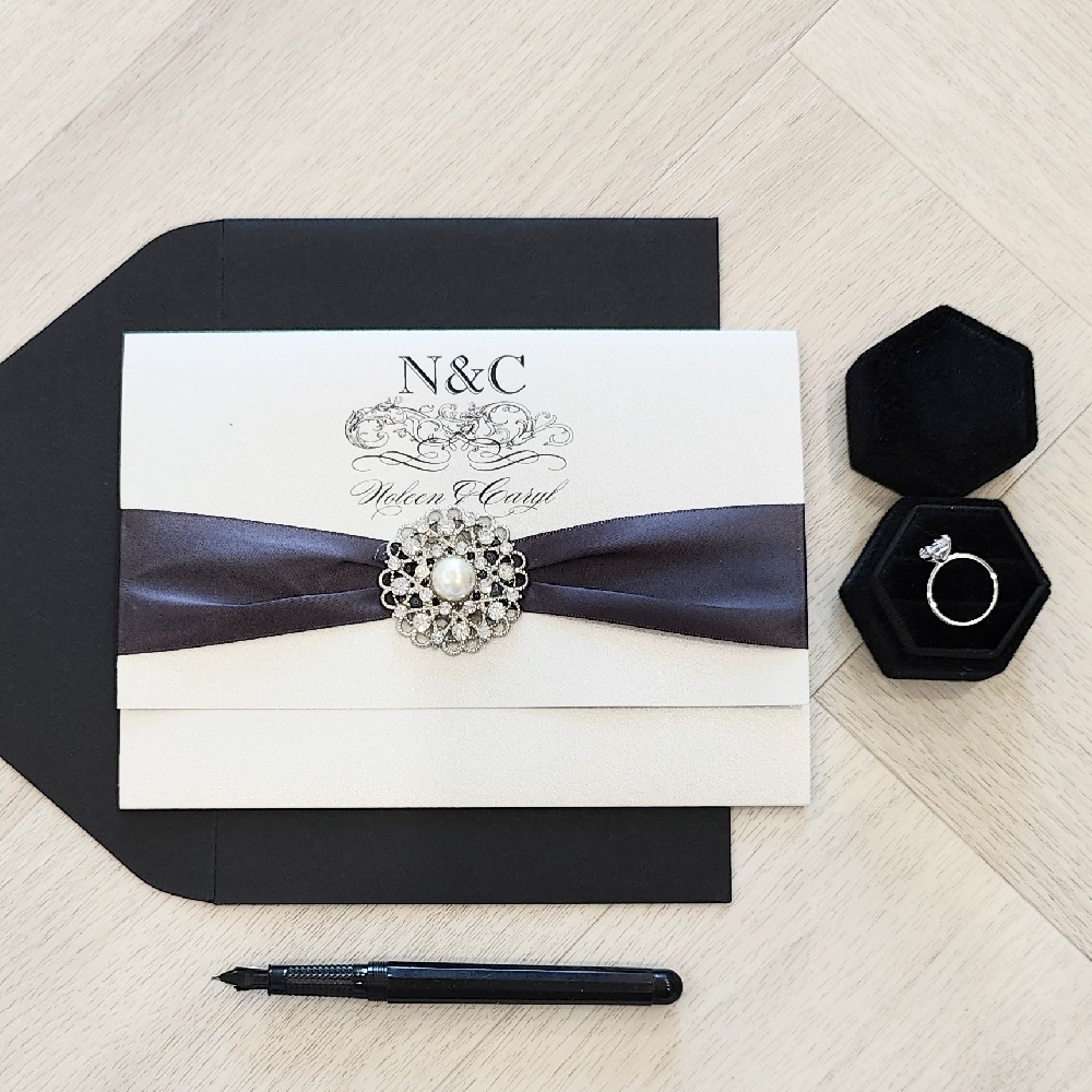 Pocketfolder style wedding invitation with a black ribbon and brooch embellishment.  It is paired with a black envelope.