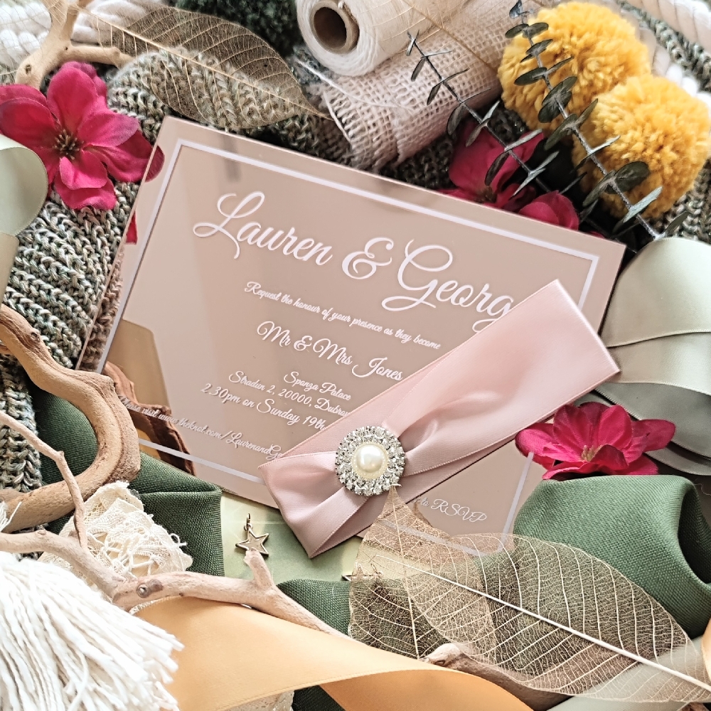 Rose gold acrylic wedding invitation with ribbon and pearl brooch.