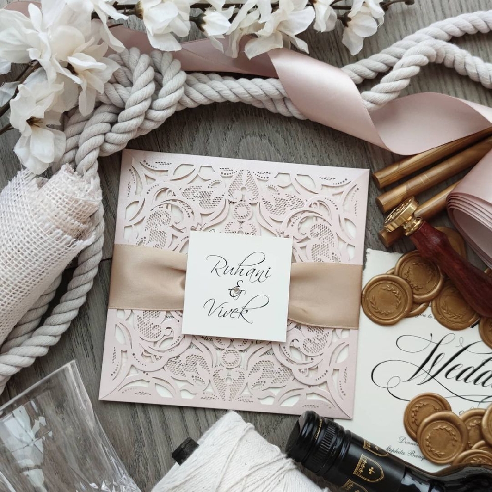 A blush colored intricately designed lasercut wedding invitation with a ribbon and tag.