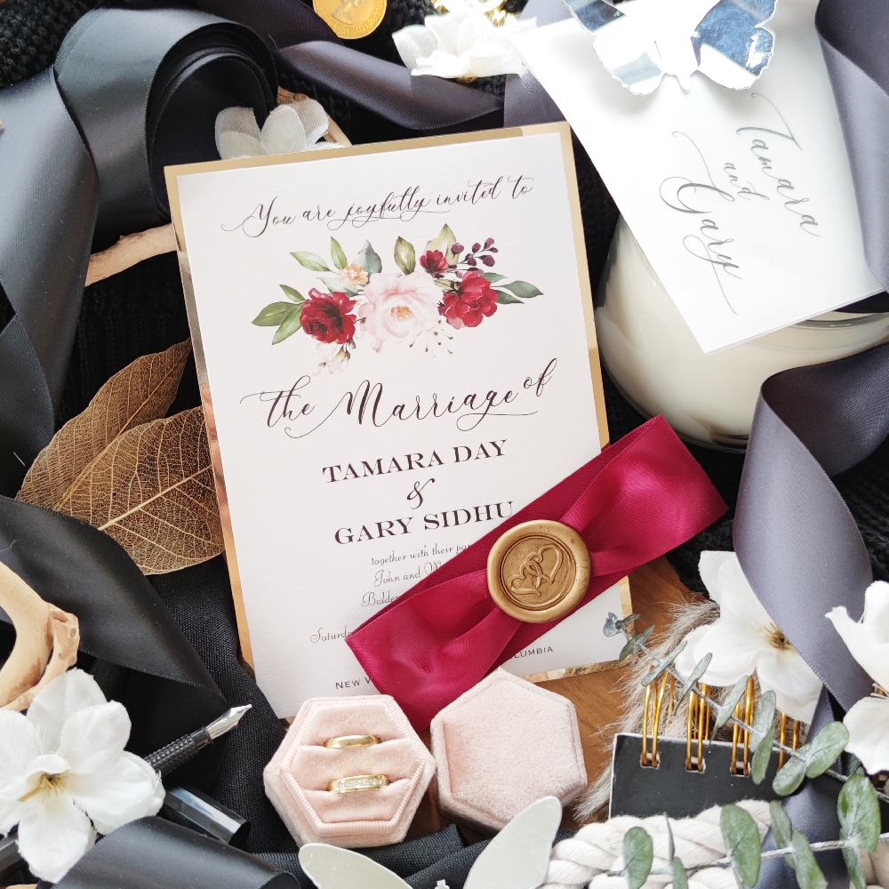 A layered card with gold chrome mirror behind ice pearl metallic paper and a deep burgundy ribbon and gold wax seal.