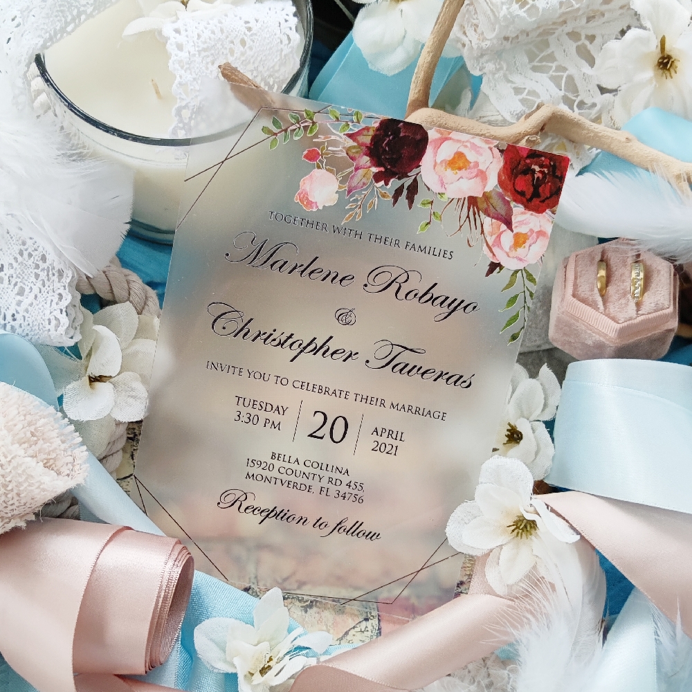 A frosted acrylic wedding invitation with red and pink flowers printed on the corner.