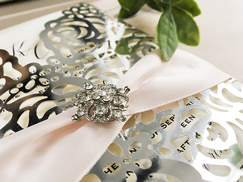 Invitation lc116: This is a silver mirror gate fold laser cut wedding invite.  The flaps have a rose design pattern.  There is a petal pink ribbon and brooch tied around.